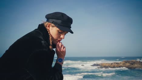 4K-UHD-Cinemagraph-of-a-young-brunette-woman-sitting-by-the-sea-with-a-black-coat-and-black-hat-with-the-waves-rushing-in-the-background