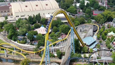 A-roller-coaster-car-of-people,-unrecognizable,-crests-the-top-of-a-roller-coaster,-then-plunges-down,-aerial-drone-view