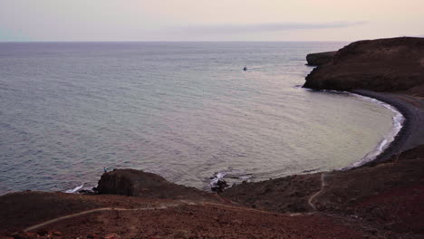Una-Toma-Estática-De-Una-Playa-Salvaje-En-Las-Islas-Canarias-De-España-Con-Un-Bote-Flotando-En-El-Horizonte-Y-Un-Hombre-Pescando-En-La-Esquina-Inferior-Izquierda