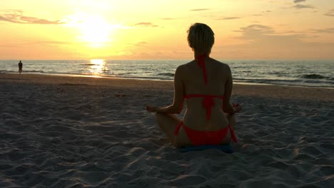 Cute-slim-woman-in-bikini-during-vacation-at-the-seaside-resort-sits-in-a-lotus-flower-position-on-a-sandy-beach-and-meditates-while-the-sun-hides-over-the-horizon