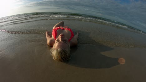 Shallow-ocean-waves-going-over-a-woman-wearing-an-orange-bikini,-using-a-fish-eye-lens