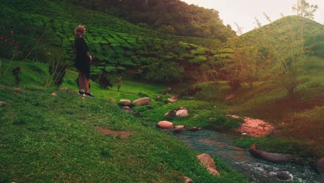 A-young-woman-is-standing-by-a-flowing-stream-near-a-tea-plantation-field-at-Cameron-Highlands-in-Malaysia