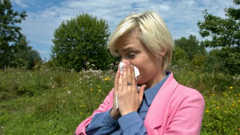 Woman-with-hay-fever-sneezing-and-blowing-into-a-tissue-whilst-in-a-green-meadow-with-flowers,-wearing-a-pink-cardigan,-blue-shirt,-blonde-hair,-light-make-up-and-green-eyes