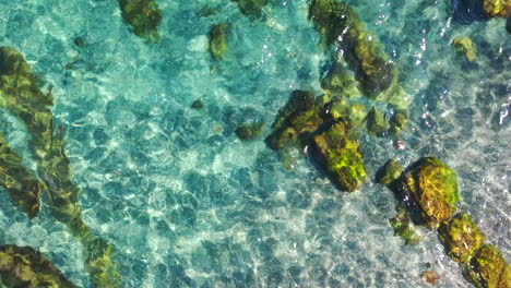 4K-Aufnahmen-Von-Oben-Nach-Unten-Vom-Kristallklaren-Mittelmeer-Mit-Felsen-Und-Sand,-Blaues-Wasser,-Das-Kleine-Wellen-Bewegt,-Urlaubsträume