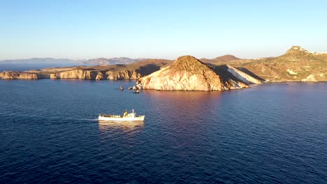 Aerial-Drone-shot-of-a-large-fishing-boat-cruising-past-the-rocky-cliff-coastline-during-sunrise-or-sunset-in-Milos-Greece