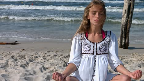 A-woman-wearing-a-white-dress,-sitting-on-the-beach-meditating,-while-beach-waves-crash-onto-the-shore