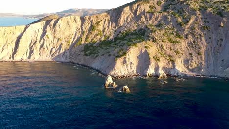 panning-aerial-drone-shot-of-deep-blue-waves-crashing-against-the-cliffs-of-a-greek-peninsula-during-golden-hour-sunrise-or-sunset