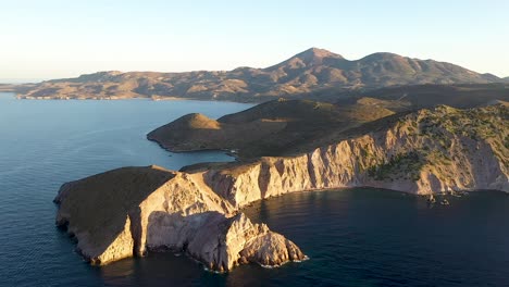 Aerial-drone-shot-of-greek-peninsula-during-golden-hour-sunrise-or-sunset-with-dramatic-cliffs-as-a-centerpiece