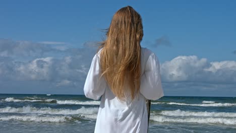 A-woman-standing-on-the-beach,-the-wind-blowing-through-her-hair-and-her-dress,-with-the-waves-crashing-in-the-background