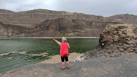 Senior,-female-pointing-to-the-cliffs-surrounding-Alkali-Lake-in-northern-Washington-state-on-a-cloudy,-breezy-afternoon