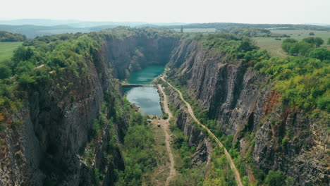 AERIAL:-Reveal-shot-and-fly-through-a-limestone-canyon-of-an-old-quarry-with-turquoise-lake-and-a-small-bridge