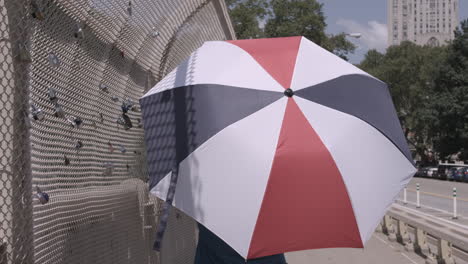 Following-shot-of-a-woman-walking-across-a-bridge-carrying-a-red-white-and-blue-umbrella-on-a-sunny-day