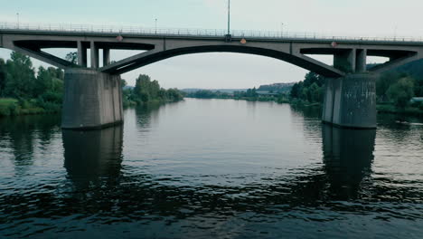 Smooth-drone-footage---flying-low-above-water-under-an-arc-bridge-over-rippled-Vltava-river-with-reflection
