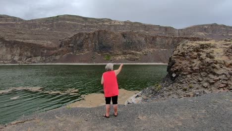 Senior,-female-pointing-to-the-cliffs-surrounding-Alkali-Lake-in-northern-Washington-state-on-a-cloudy,-breezy-afternoon