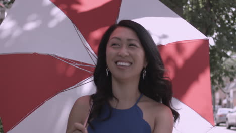 Happy-Asian-woman-strolls-down-a-city-street-carrying-a-red-and-white-parasol-on-a-sunny-day