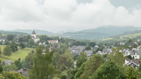 Time-lapse-of-a-valley-in-Sauerland,-Germany,-with-clouds-passing-by-the-village-of-Grafschaft-with-a-clear-view-of-the-monastery-and-typical-half-timbered-houses-against-a-sloping-mountain-landscape