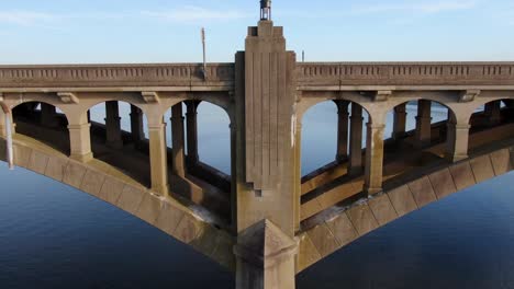 Langsame-Absteigende-Luftaufnahmen-Zeigen-Eine-Schöne-Betonbrückenarchitektur-Und-Ein-Designmuster,-Das-Ruhiges-Blaues-Flusswasser-Enthüllt