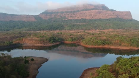 taking-off-Early-morning-as-soon-as-the-first-rays-of-sun-hits-the-foothills-of-Sahyadri-ranges-near-Lonavala,-India---Aerial-drone-footage-with-lake