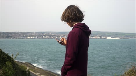 Junger-Teenager-Junge,-Der-Auf-Seinem-Handy-Soziale-Medien-Mit-Blick-Auf-Die-Küste-Im-Hintergrund-Durchsucht,-Verschiedene-POV