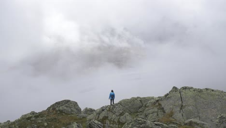 A-lone-female-hiker-stands-on-the-edge-of-a-rocky-outcrop-looking-out-over-a-large-alpine-glacier-as-the-fog-and-mists-swirl-around-her