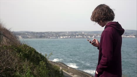 Young-teenager-boy-searching-social-media-on-his-mobile-phone-with-coastal-view-in-background,-copy-space