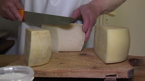 Cheese-maker-in-white-apron-cuts-piece-of-local-cheese-with-giant-knife,-offers-in-to-customer,-closeup-static-shot