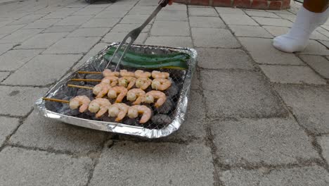 Locked-off-view-of-disposable-mini-BBQ-grill-with-Zucchini-and-Shrimp-skewers-male-hand-poking-and-turning-around-food