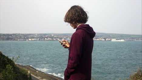 Junger-Teenager-Junge,-Der-Auf-Seinem-Handy-Soziale-Medien-Mit-Blick-Auf-Die-Küste-Im-Hintergrund-Durchsucht,-Verschiedene-POV