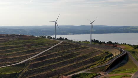 Aerial-turn-reveals-landfill-terraces-on-hillside,-high-atop-Susquehanna-River,-wind-turbines-rotating-on-summer-day