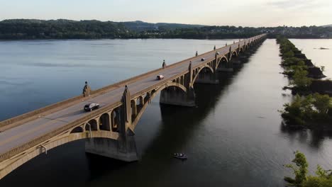Aerial-tracking-traffic-drone-shot-of-Susquehanna-River-bridge-at-sunset-fishing-boat-on-water,-former-bridge-stone-piers-covered-with-vegetation-and-trees