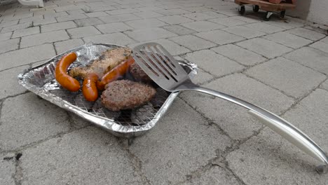 Locked-off-view-of-disposable-mini-BBQ-grill-with-different-meats-and-spatula-leaning-against-it