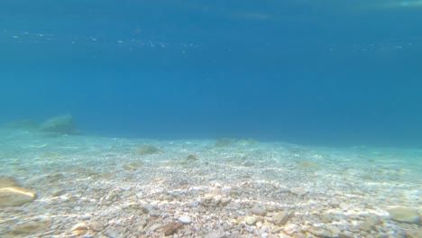 Underwater-footage-of-a-seabed-with-white-pebbles,-blue-sea-and-reflections-on-the-surface
