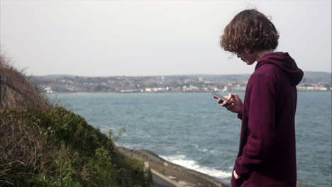 Junger-Teenager-Junge,-Der-Soziale-Medien-Auf-Seinem-Handy-Mit-Blick-Auf-Die-Küste-Im-Hintergrund-Sucht,-Kopierraum