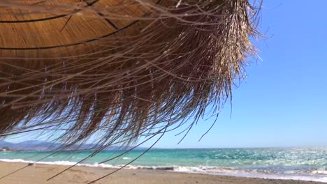 Strohgedeckter-Tiki-regenschirm-An-Einem-Windigen-Strand-Mit-Meerblick-In-Tropischer-Lage,-Marbella-Malaga-Spanien