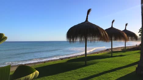 Dreamy-paradise-vacation-shot-moving-to-the-right-on-Marbella-Malaga-sea-view-in-Costa-del-Sol-in-Spain,-green-tropical-plants-and-thatched-tiki-umbrellas-at-the-beach