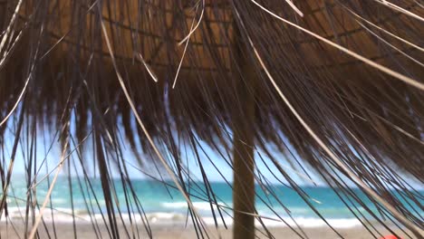 Wind-blowing-through-thatch-tiki-umbrella-at-the-beach-in-Marbella-Malaga-Spain,-tropical-holiday-vibes