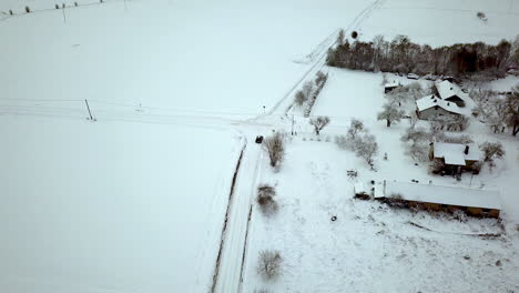 Slow-aerial-following-shot-of-car-driving-away-on-frozen-and-snow-covered-rural-road-next-to-snow-covered-rooftops