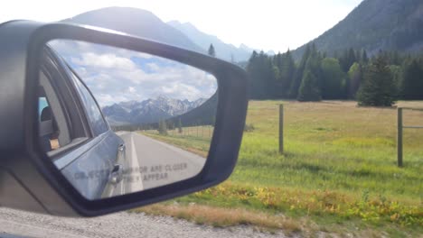 Landscape-view-of-the-side-mirror`s-car-while-moving-with-reflection-view-of-the-highway-and-surrounding-with-green-pine-tree-forest-and-rockies-mountain-in-Alberta,Canada