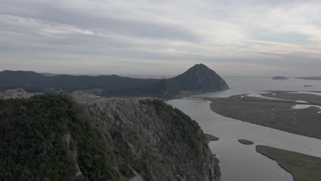 Landscape-view-of-mountain-with-abandoned-quarry-on-its-summit-and-reveal-of-river-estuary,-the-mountain-ridge-in-the-distance-with-ships-anchored-in-the-bay,-Russia