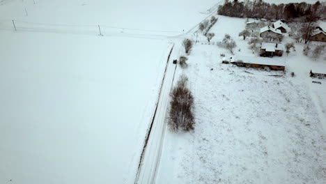 Slow-aerial-following-shot-of-car-driving-and-reaching-intersection-on-frozen-and-snow-covered-rural-road-next-to-snow-covered-rooftops-and-then-turning-right-to-residential-area