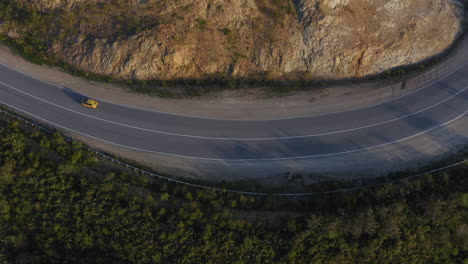Top-view-of-a-yellow-color-car-passing-on-a-long-curved-road-in-the-green-mountains-with-a-cliff-on-it's-right-side,-during-sunset