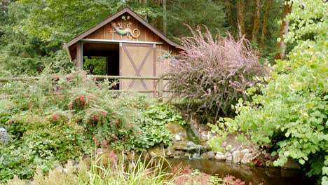 Beautiful-and-peaceful-wooden-barn-and-water-pond-in-a-wooded-forest-country-setting-with-blooming-flowers-and-plants-surrounding-the-area