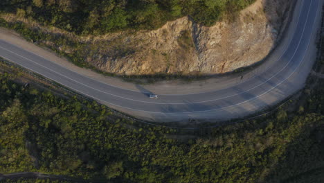 Top-view-of-a-white-color-car-passing-on-a-long-curved-road-in-the-green-mountains-with-a-cliff-on-it's-right-side,-during-sunset