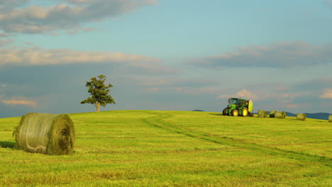 Landwirtschaftlicher-Traktor,-Der-Tagsüber-Heu-Unter-Einem-Hügel-Mit-Einem-Verlassenen-Baum-Entlädt,-Mit-Blick-Auf-Einen-Heuhaufen-Und-Blick-Auf-Sich-Bewegende-Wolken-Und-Die-Umliegende-Natur-&#39;Landschaftsansicht-4k