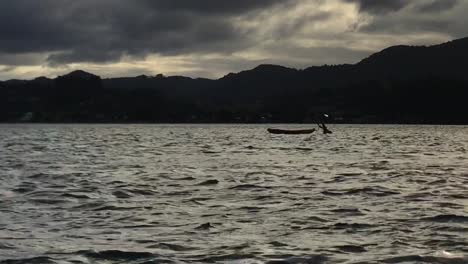 Kayak-floats-gently-on-the-surface-of-a-rippling-lake-at-sunset-against-a-mountainous-backdrop-while-a-bird-in-silhouette-flies-across