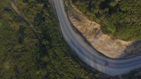 Top-view-of-a-red-color-car-passing-on-a-long-curved-road-in-the-green-mountains-with-a-cliff-on-the-left-side,-during-sunset