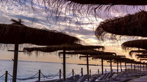 4k-sunset-in-paradise-beach-of-Marbella,-beautiful-purple-pink-clouds-moving-and-view-of-sunbeds-and-tropical-straw-umbrellas