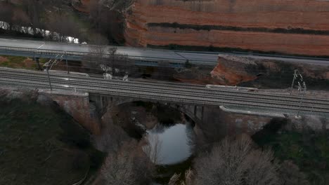 Aerial-shot-moving-backwards-from-a-railway-over-a-river-with-a-train-passing-through-it,-near-a-traffic-road-and-a-dirt-road