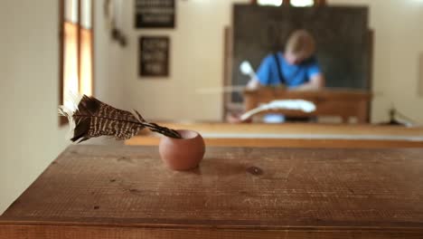 Female-teacher-or-student-in-soft-focus-writing-notes-in-classroom-with-feather-quill-ink-pen