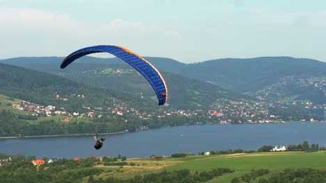 A-single-paraglider-makes-long-turn-right-against-forested-mountains-with-small-buildings-and-vast-lake